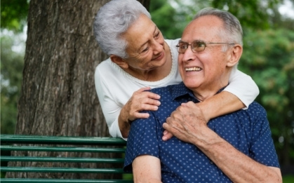 Elderly women leaning forward to embrace an elderly man sitting on a park bench