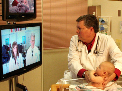 A doctor holds a baby to while speaking to two other doctors on a screen via video.