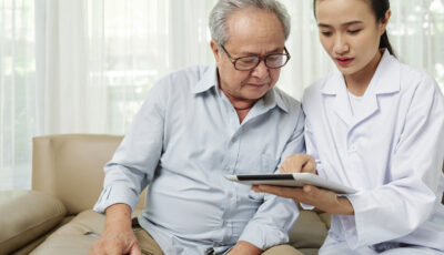 An elderly man wearing glasses is sitting next to a younger woman in a lab coat, who is holding a tablet. He is looking at the tablet that is in her hand. 