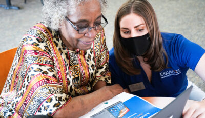 An elderly woman is on the left of the screen, and a younger woman wearing blue scrubs and a mask is sat next to her, showing her something on a laptop, which is placed on a table. 