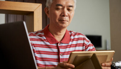 An older man in a red striped polo is sitting at a desk and looking at a framed photo. 
