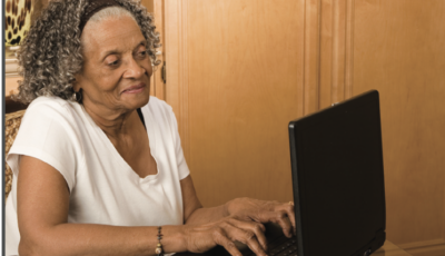 An elderly woman in a white t-shirt is sitting and typing on a black laptop.