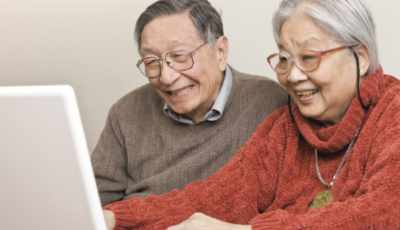 An older man and woman are sitting and looking at a white tablet on the left side of the picture. Both are wearing glasses. 