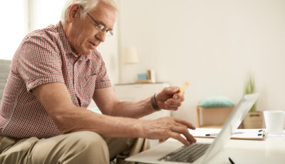 An older man wearing glasses is sat on a couch, and is typing something on the laptop on the coffee table in front of him. 