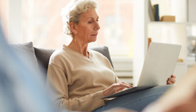 An older woman is sitting on a couch, a laptop open on her lap. She is looking at the laptop.
