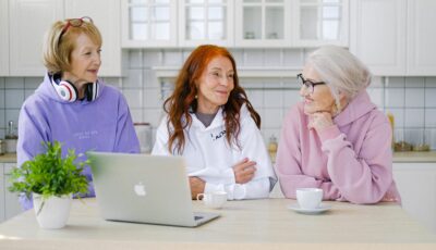 Three women are sitting at a table. From left to right, there is a woman with a purple hoodie and headphones around her neck, a woman with a white hoodie on and red hair, and a woman with a pink hoodie on and glasses. On the table in front of them is a potted plant, an open laptop, and two coffee cups.