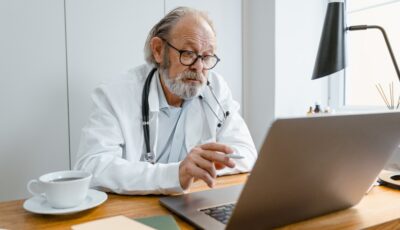 An older man with glasses, a white lab coat, and a stethoscope around his neck is sitting at a desk looking at his open laptop. There is a cup of coffee on his right on the desk.