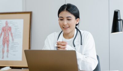 A woman in a lab coat and with a stethoscope around her neck is sat at a desk, looking at a laptop in front of her. 