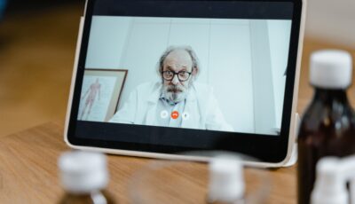 A tablet is sat on a table, and an older man in a lab coat and glasses is seen on the screen. He is looking directly at the camera. 