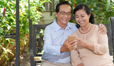 A couple is sitting next to each other outside. The man on the left is holding a phone, and the woman on his right is pointing at something on the phone. Both are smiling. 
